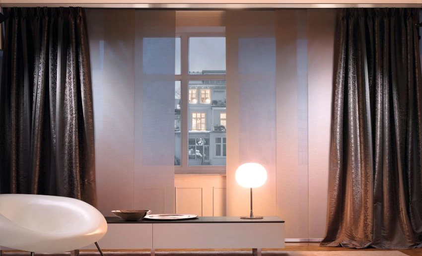 Dunkelbraune blickdichte Flächenvorhänge vor einem abendlichen Fenster in einem schwach beleuchteten Raum